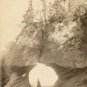 Black and white photograph of arched rock formation with woman in long dress standing in open arch on beach