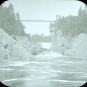 Black and white image of river, rocky shoreline and suspension bridge in the background